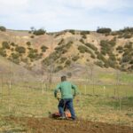 A farmer working with a tiller in an open countryside field, preparing the soil for planting.